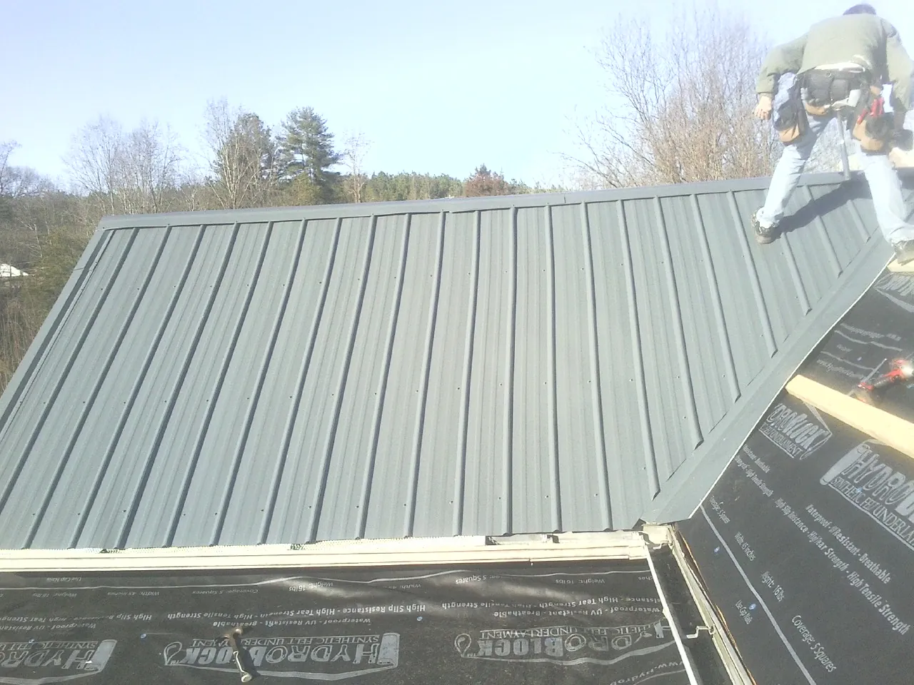 Crew installing dark green standing seam panels over synthetic underlayment in Putnam Station
