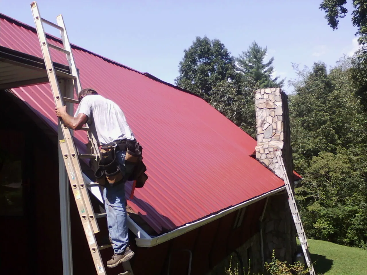 Roofer on a ladder beside a red metal roof and stone chimney — Soffit & Fascia Repair by Roofing Brothers in Putnam Station